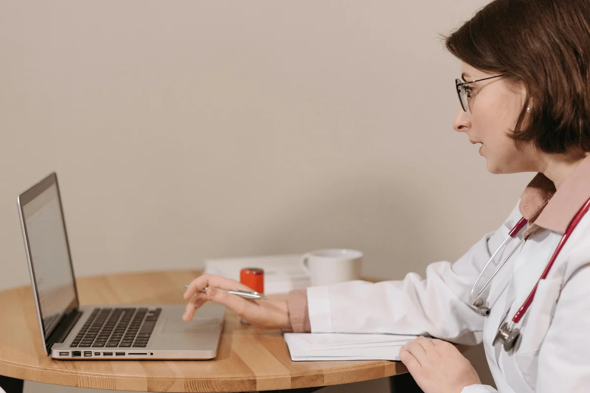 Female doctor using a laptop at a desk with notebook and pen in a medical office.