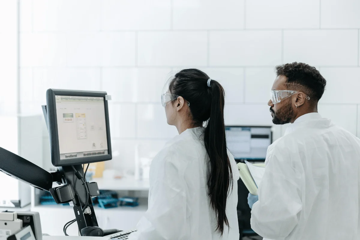 Two scientists in lab coats and safety goggles work in a laboratory, focusing on a computer screen displaying data.