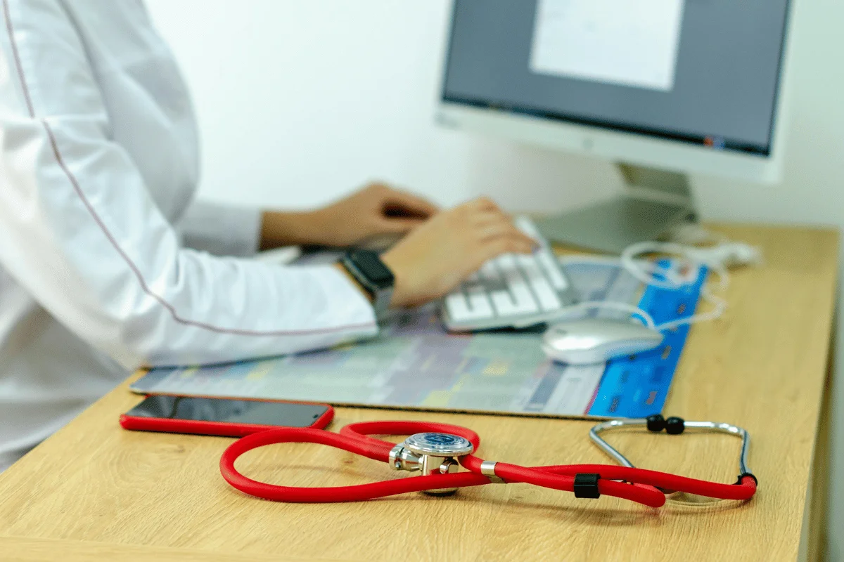 A doctor types on a keyboard at a desk with a red stethoscope, smartphone, and computer mouse.
