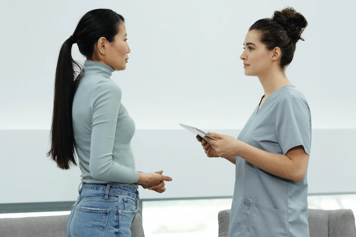  nurse in scrubs speaks with a woman in jeans and a turtleneck, holding a tablet.