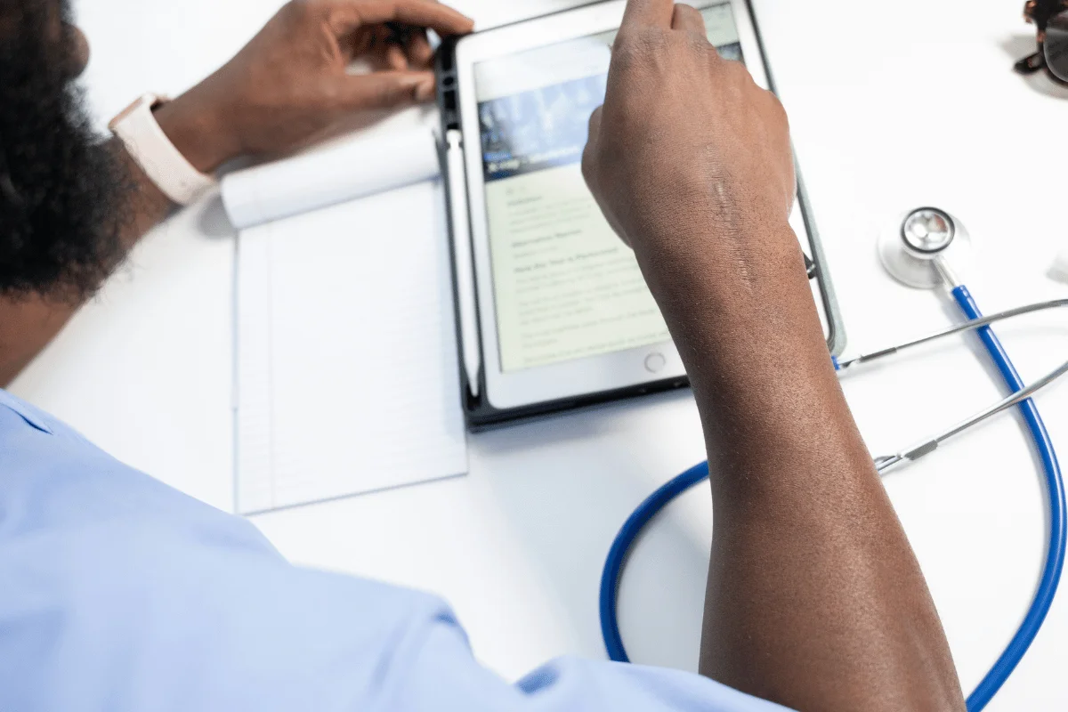 A person in medical attire reviews information on a tablet, next to a notebook and stethoscope on a white desk.