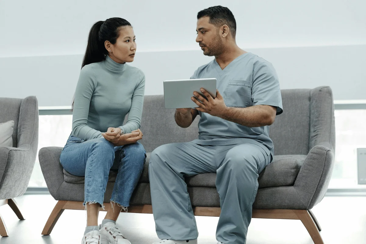 A doctor in scrubs sits on a gray couch showing a tablet to a woman in a teal sweater and jeans.