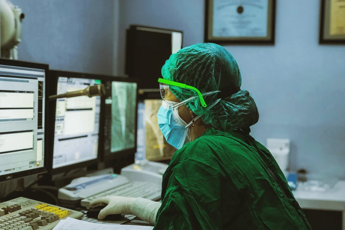 A healthcare worker in green scrubs and protective gear operates multiple computer monitors, focused and attentive, in a clinical setting.