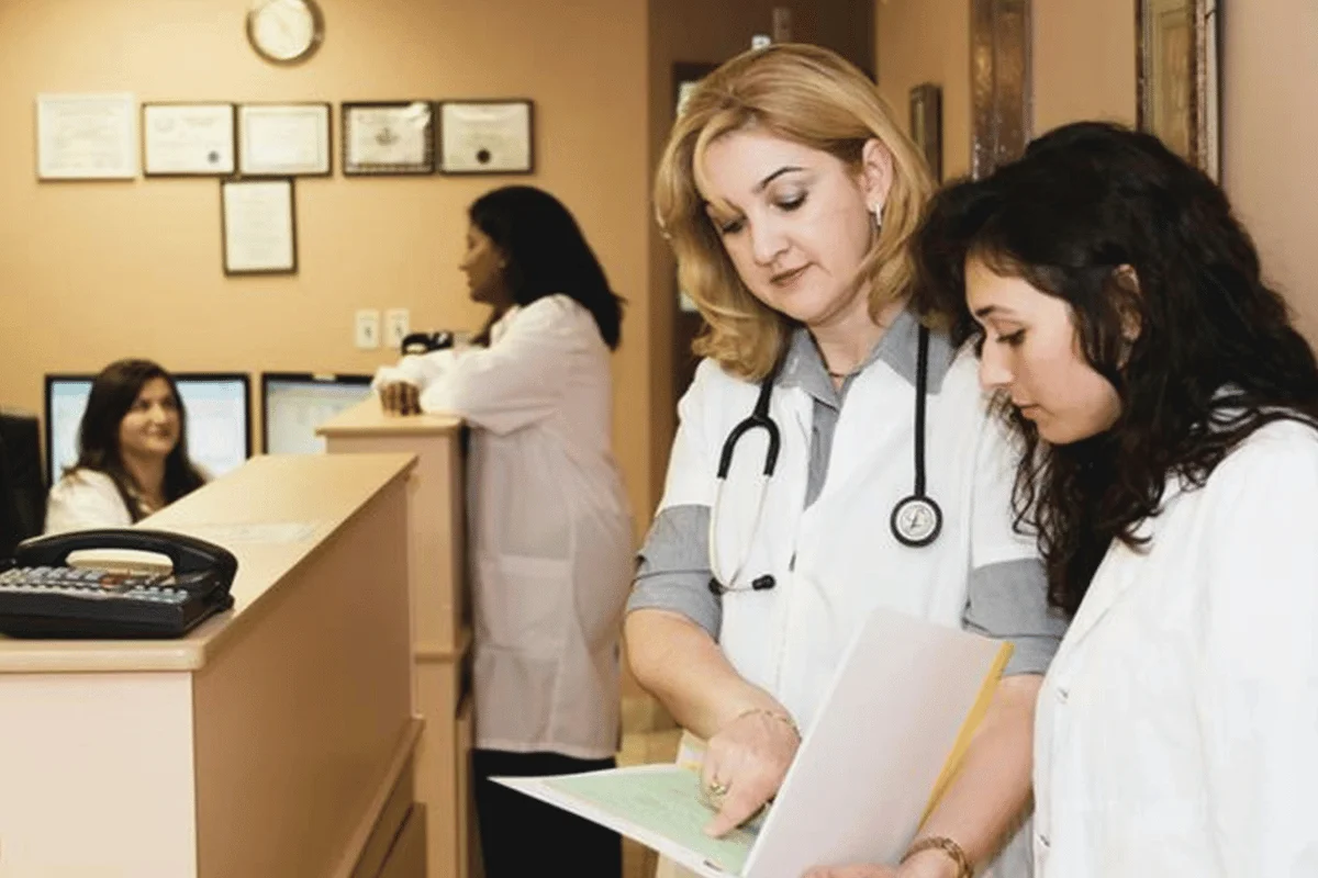 Doctors in a clinic are reviewing patient files. A woman in a lab coat and stethoscope consults a colleague.