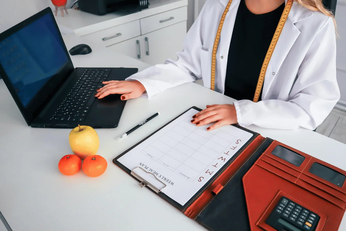 A person in a lab coat sits at a desk with a laptop and planner. A measuring tape is draped over their shoulders.