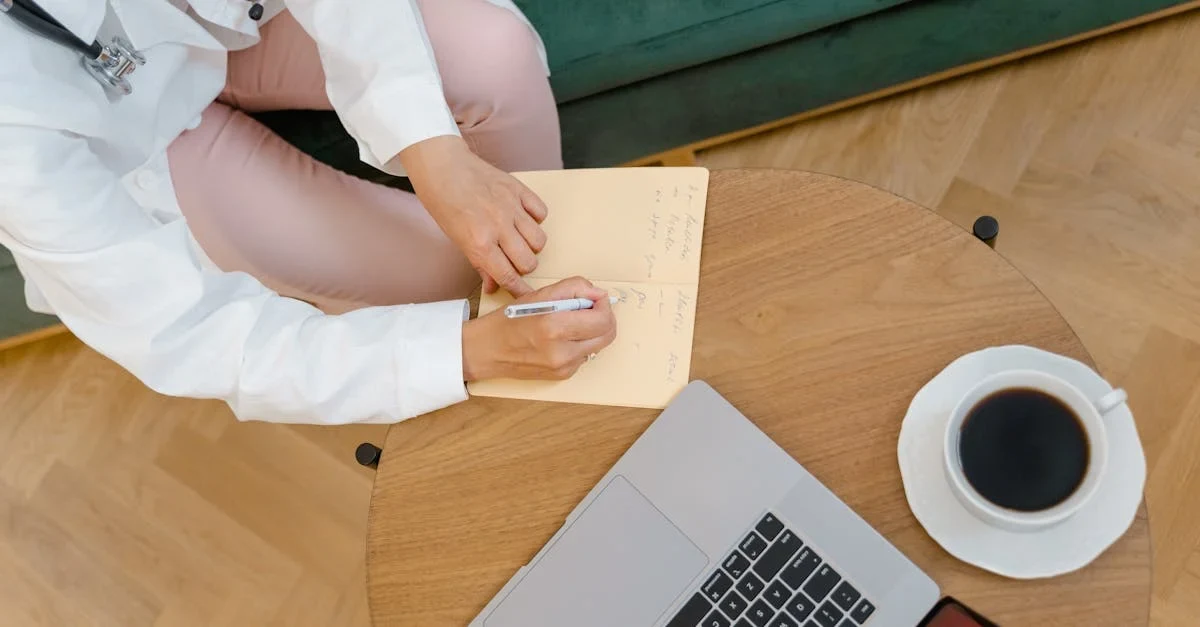 Healthcare professional using laptop and mobile device to implement autonomous medical coding software at wooden desk.