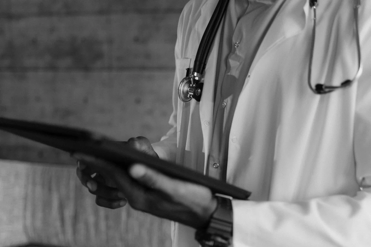 Black and white image of a doctor in a lab coat with a stethoscope, holding and reading from a tablet. 