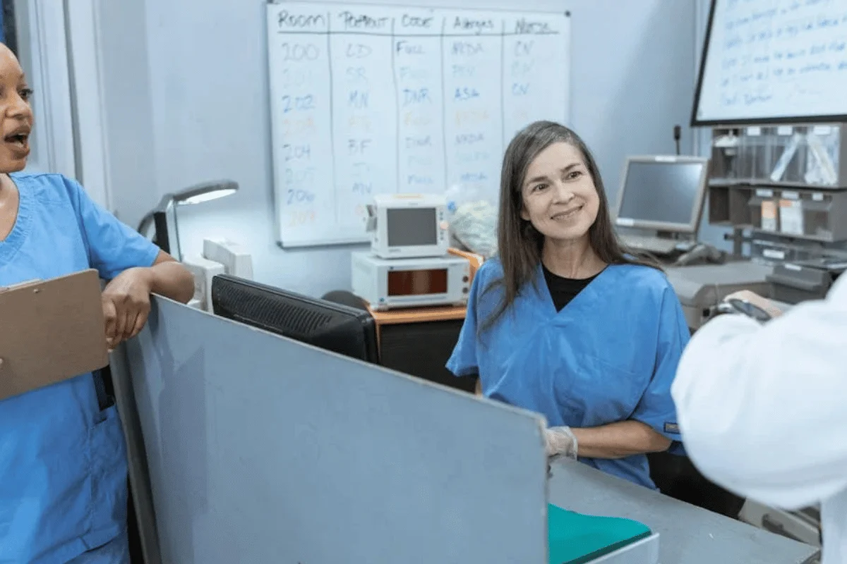 Medical staff in blue scrubs working at healthcare facility reception desk with computer and scheduling charts.