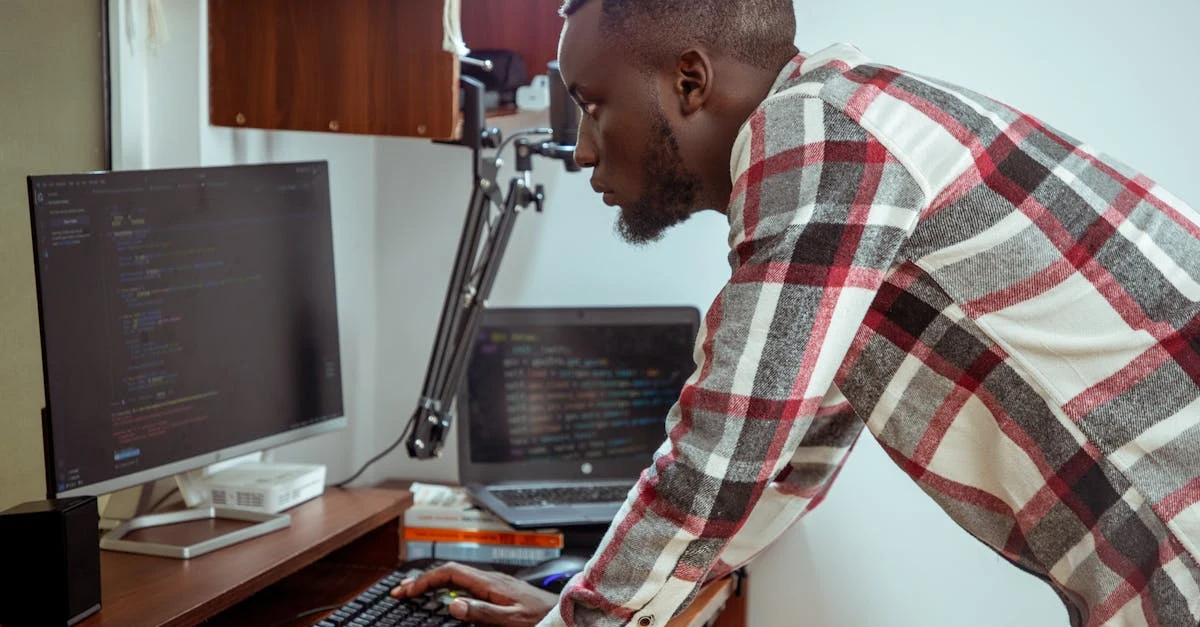 Developer in plaid shirt coding ambient AI scribe software on dual monitor setup at wooden desk.