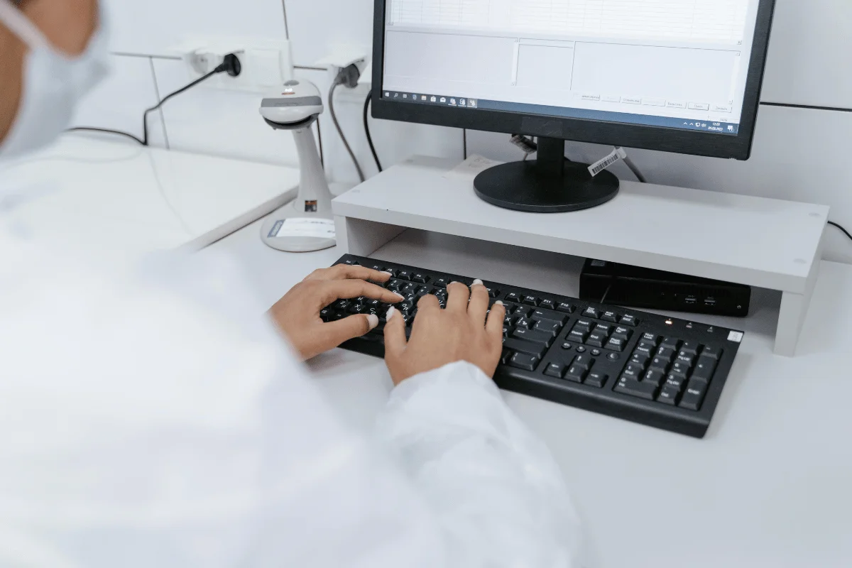 Person in white lab coat typing on a keyboard in front of a computer screen displaying data, in a sterile lab setting.