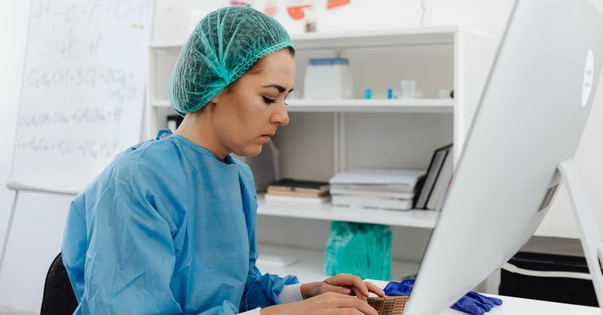 Healthcare professional in surgical attire using computer for spine surgery coding and medical documentation at workspace.
