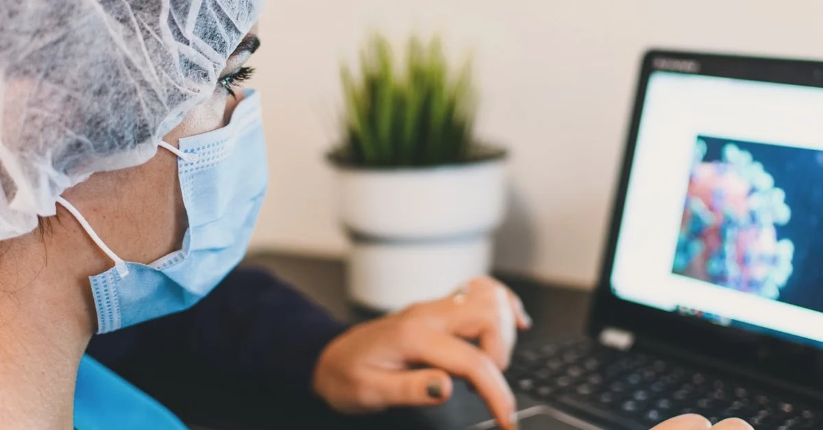 Healthcare professional reviewing ambulatory surgery coding guidelines on laptop while wearing protective gear.