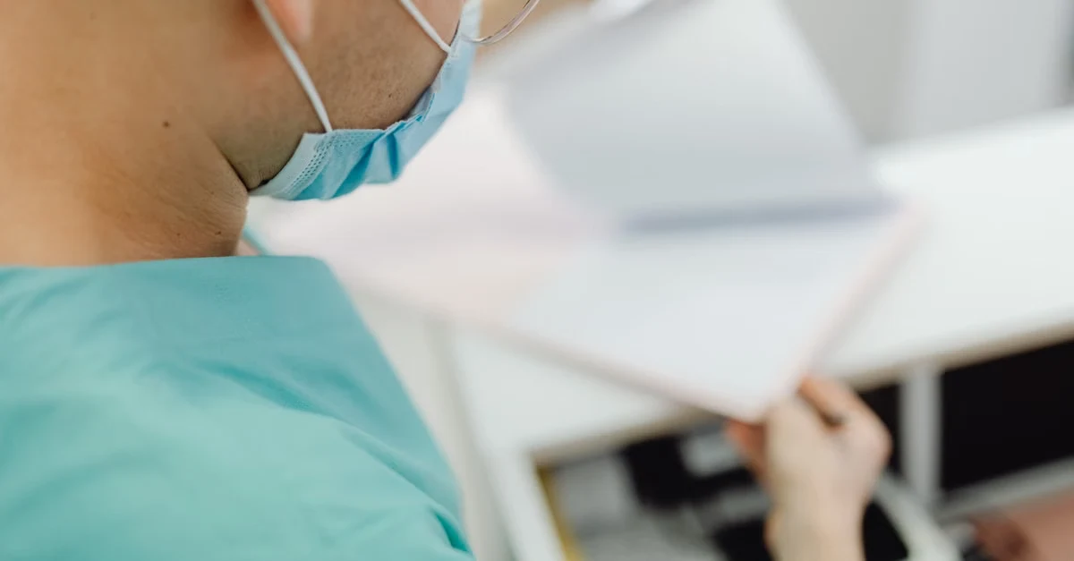 Surgeon in teal scrubs reviewing hand surgery coding documents during medical procedure consultation.