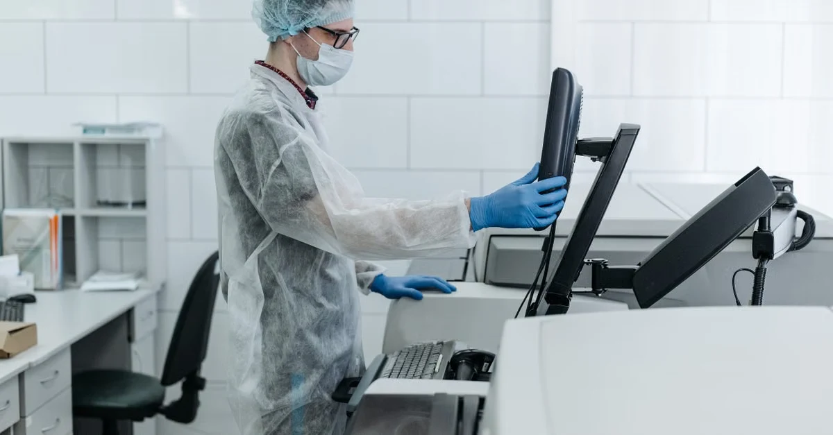 Healthcare worker in sterile protective gear disinfecting computer workstation in clinical office environment.