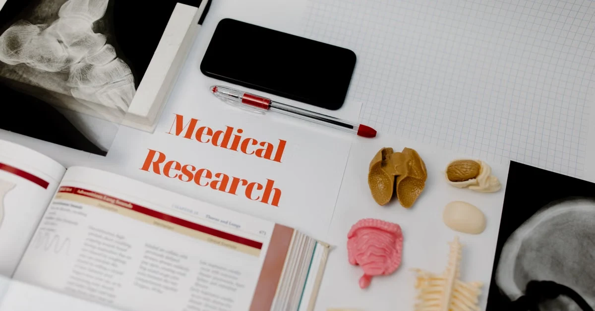 Flat lay of medical research materials including anatomical models, x-ray images, smartphone, and pen on white desk.
