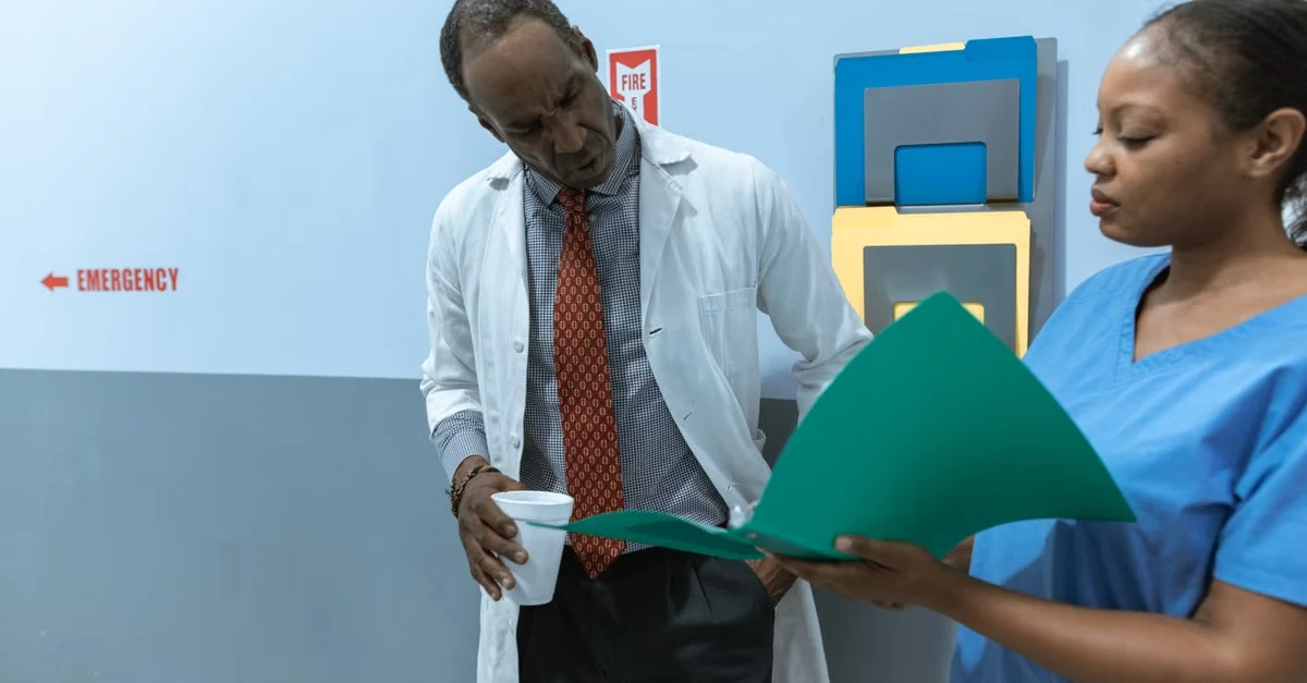 Doctor and nurse review a green medical folder in a hospital hallway, with emergency signage behind them, conveying a focused, professional tone.
