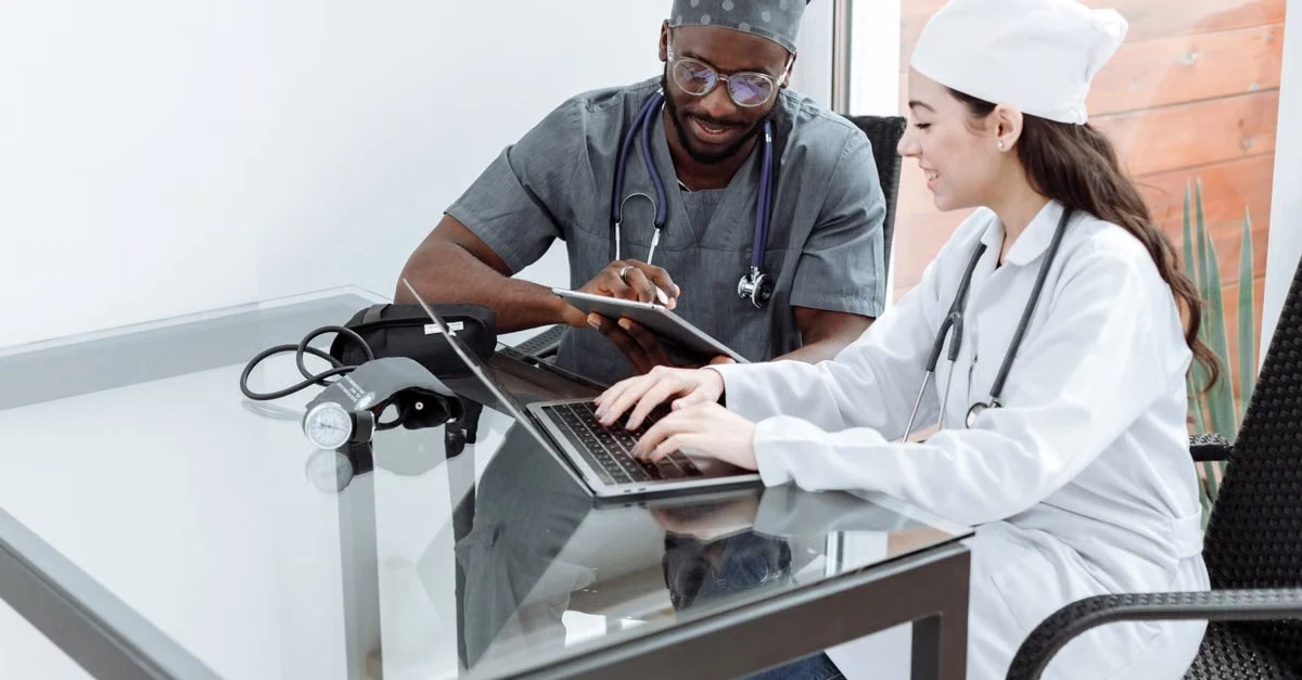 Two healthcare professionals reviewing medical records on laptop at clinical desk during consultation.