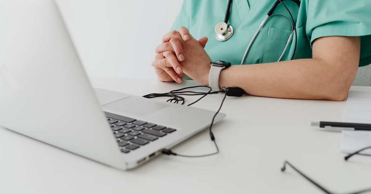 Healthcare professional in green scrubs sits at a desk with clasped hands, stethoscope, laptop, and medical tools, conveying a calm clinical setting.