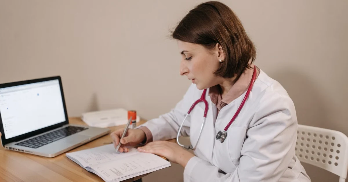 Doctor in white coat writing patient notes at desk with laptop, demonstrating ambient clinical documentation workflow.