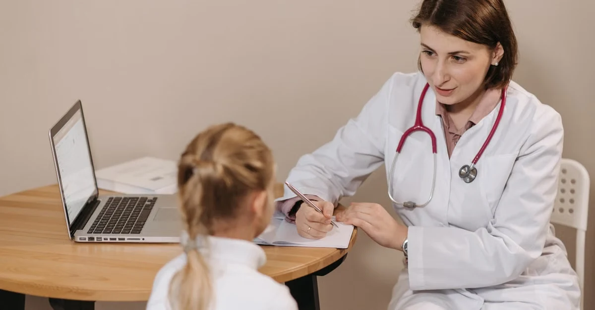 Female physician documenting medical coding risk adjustment notes during patient consultation at desk with laptop.