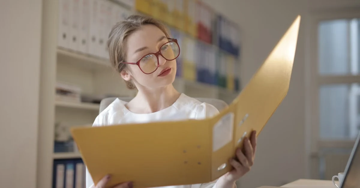 Woman reviewing healthcare charge capture documentation in bright office with shelves and laptop visible behind her.