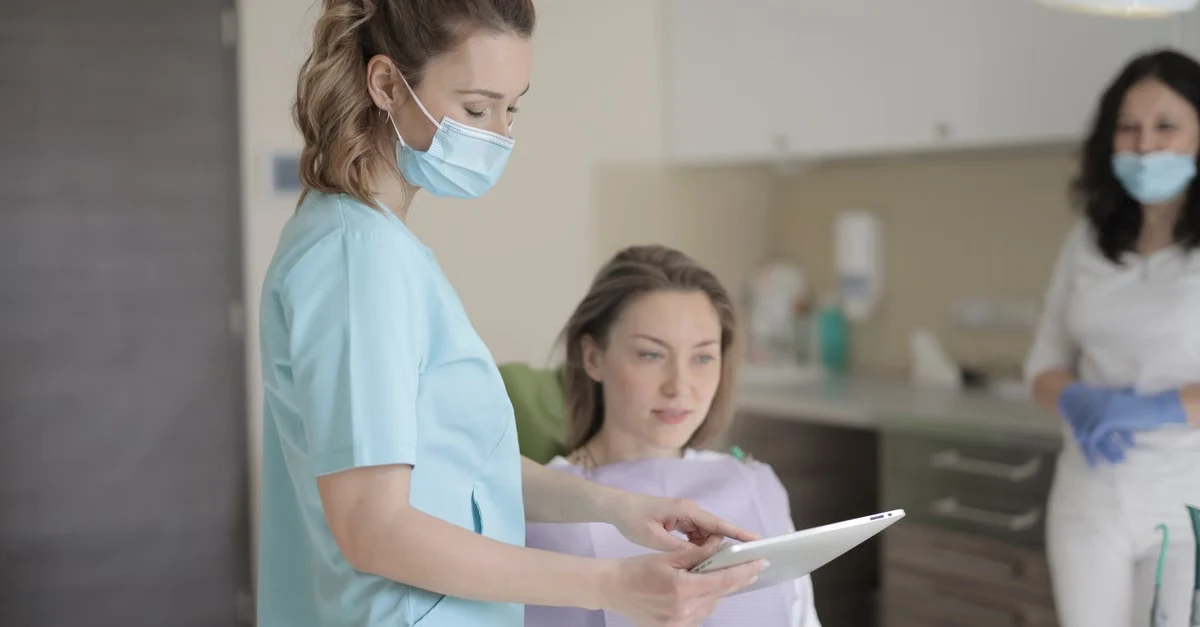 Healthcare professional in blue scrubs reviewing digital documentation with patient at clinic reception desk.