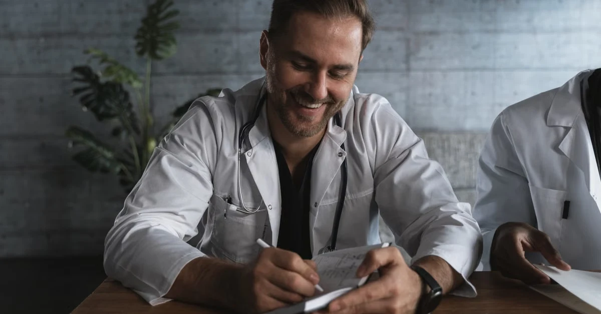 Male physician in white coat with stethoscope documenting patient notes for HCC medical coding requirements.
