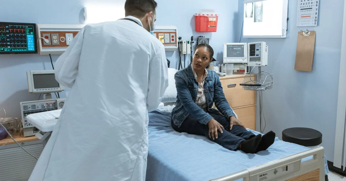 Doctor examines patient on emergency room bed with medical equipment and monitors in background during clinical assessment.