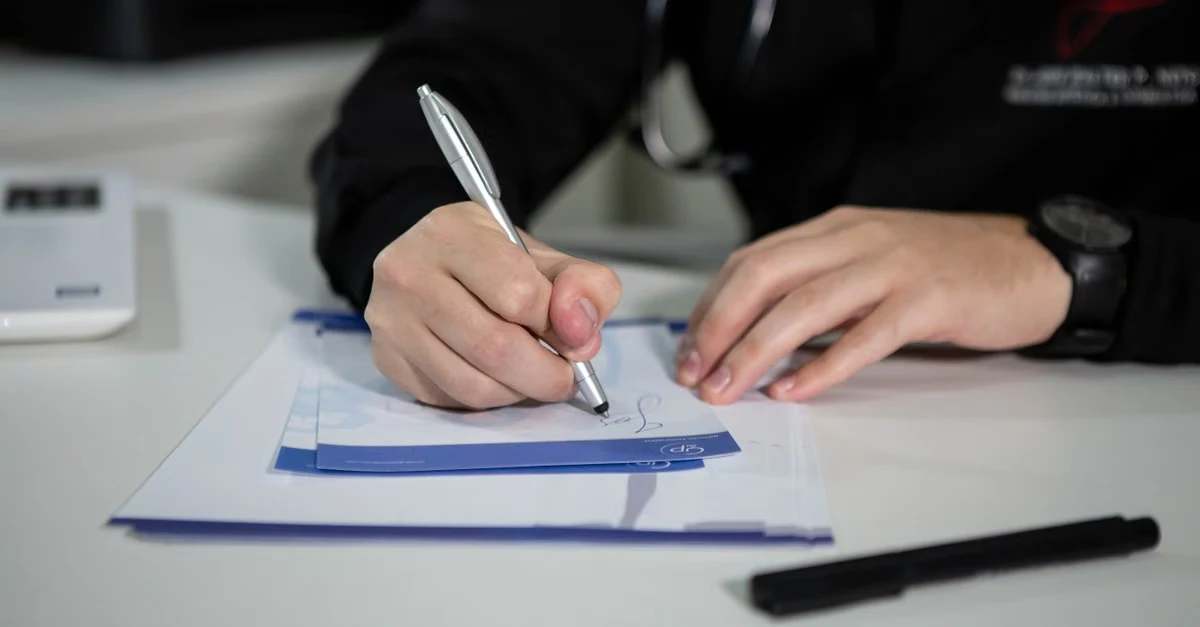 Person writing on medical form at desk with pen and clipboard during patient documentation.