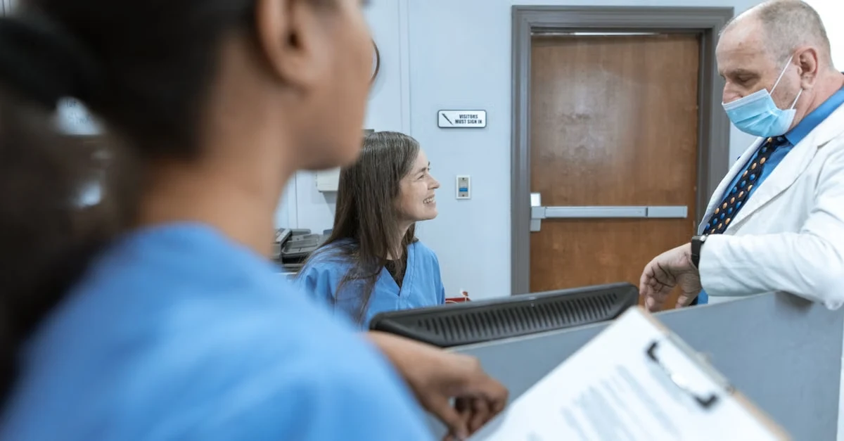 Doctor with face mask discussing patient chart with healthcare staff at clinic reception desk.