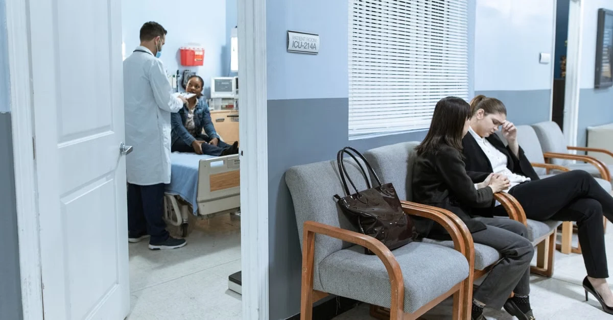 Patients waiting in modern medical clinic examination room while doctor consults with family inside.