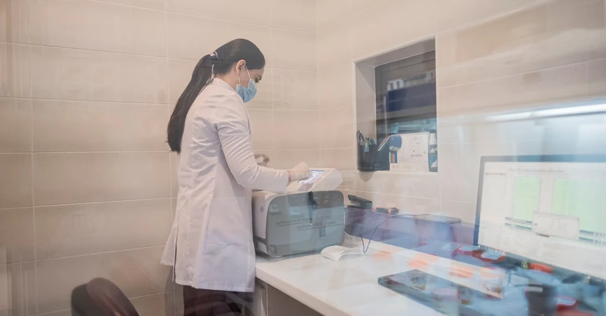 Female cardiologist in white coat using AI scribe cardiology technology at computer workstation in medical office.