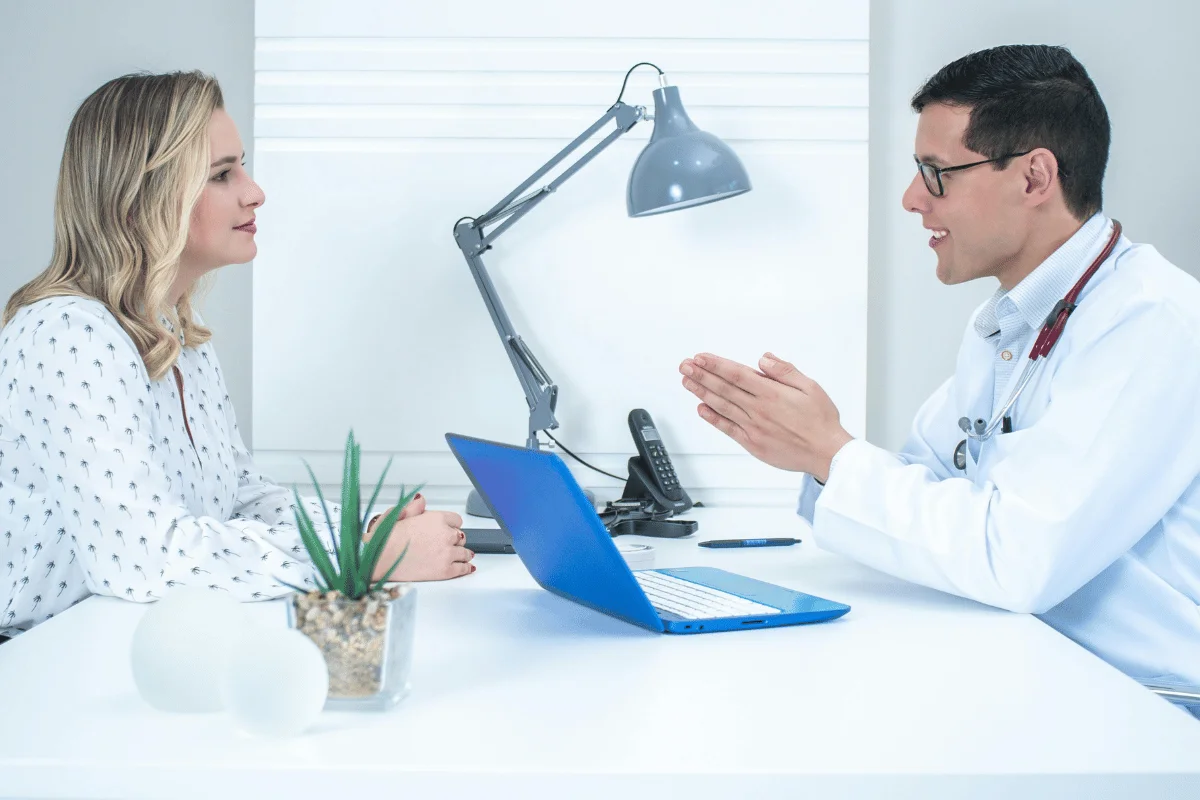 A doctor in a white coat talks to a woman at a desk with a blue laptop and a plant.