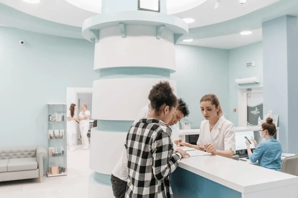 A modern medical reception area with calming light blue walls features two patients speaking with a receptionist at the front desk, creating a bright, welcoming healthcare environment.