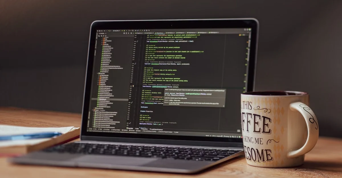 A laptop screen displaying code in a dark-themed editor sits on a wooden desk next to a ceramic mug.
